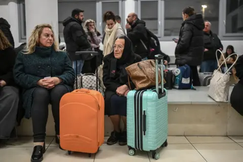 Gambar Getty Dua wanita duduk di bangku di terminal feri. Mereka berdua memiliki koper dan tas dan tidak tersenyum. Di latar belakang ada banyak orang lain yang terbungkus tas hangat dan membawa.