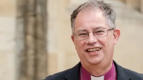 Diocese of Oxford Bishop Steven smiling at the camera has grey hair, is wearing glasses and has a purple clerical shirt.