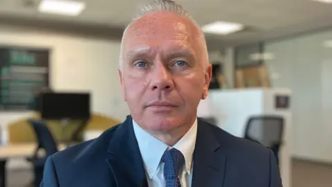 Alex Dunlop/BBC A man with short grey hair looks directly into the camera. He sits in a meeting room which has been blurred behind him. He wears a black suit jacket with a white shirt and blue tie. 