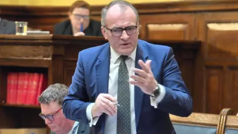 BBC Alfred Cannan, a man who wears a navy suit, he has a spotted tie, and looks serious, he wears glasses and holds his hand up in the House of Keys, there are wooden panels behind.