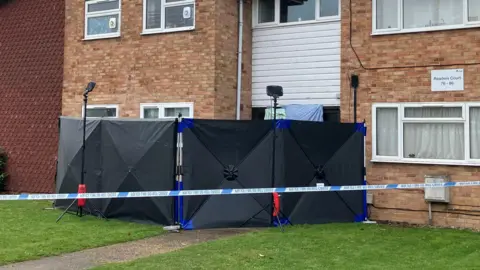 Lewis Adams/BBC Black screens and police tape blocking off the entrance to a two-storey block of flats.