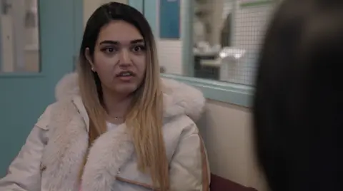 A woman with long hair wearing a pale coat with a fur-style collar looks at someone off camera.