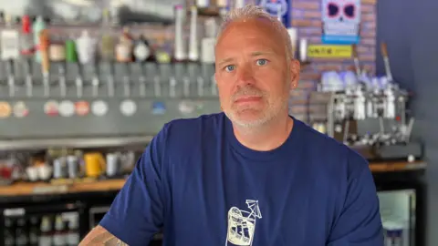 Shariqua Ahmed/BBC Neil Treliving, owner of the Blind Tiger, in front of a bar, optics behind him. He wears a blue-T-shirt.