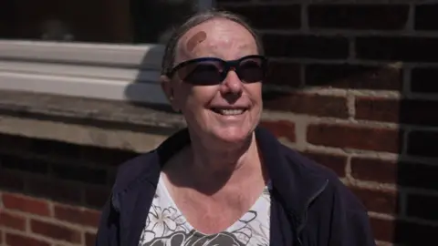 Glynis Nifton, wearing a white, black and beige floral top, a navy zip-up fleece and black sunglasses, smiling at the camera. She has brown hair tied back and a small plaster on her forehead. She is stood in the street in front of a red brick building.