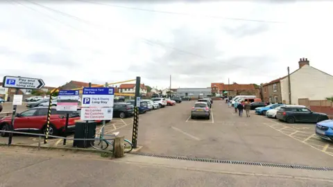 Google The entrance to a full surface car park. There are clouds above. There is a blue bicycle leaning against a sign in the foreground.