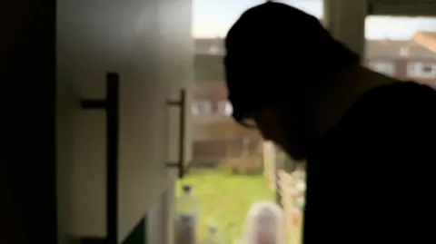 A man whose face is not visible is on the right of the photo. Cupboards are on the left and a garden is in the middle.