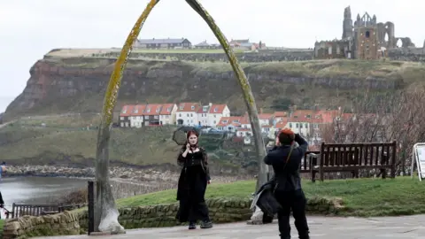 Reuters A man takes a pictures of a woman wearing all black standing underneath the whale bones on Whitby seafront - the abbey is visible in the background