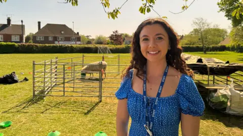 George Carden/BBC Kerry Saunders looks at the camera and smiles. She is wearing a blue top and has long brown hair. In the background there is a sheep pen with a sheep in it on a school field.