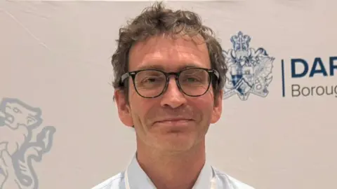Michael Walker with light brown hair and black glasses standing in front of board which says "Darlington Borough Council". He is wearing a shirt.