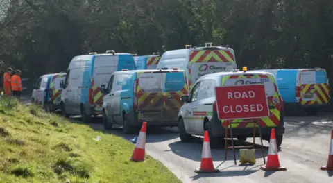 A number of South East Water vans behind each other in front of a sign advertising a road closure. There are four cones in front of the road closed sign. 