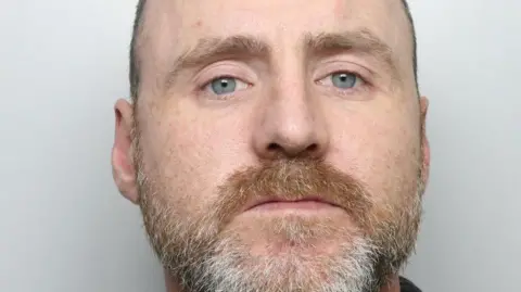 A man staring straight ahead. He has a ginger and grey-coloured beard with very short hair.