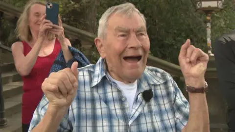 George has his hands in the air and is cheering. He is wearing a check shirtand has grey hair.