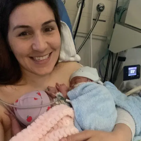 Lowri Price Lowri Price holds her baby twins in a hospital neonatal care unit. One infant wrapped in a pink blanket and another in a light blue blanket.