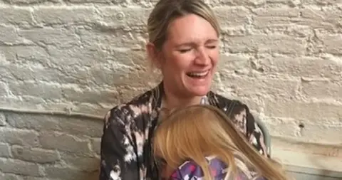 Geraint John Deb holding her young daughter who is cuddled into her. Deb is laughing and has her blonde hair toed back. She is wearing long sleeved tie-dye effect top in grey white and pink. Her daughter has long blonde hair. 