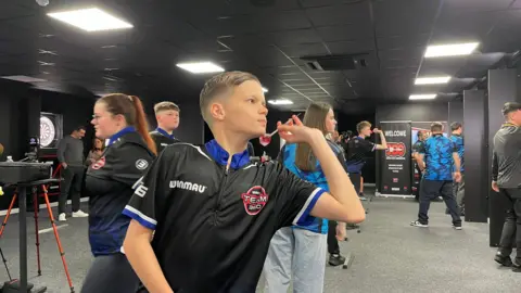 Jack throws a dart at a board at the new Hangar61 facility. There are lots of other young players around him.