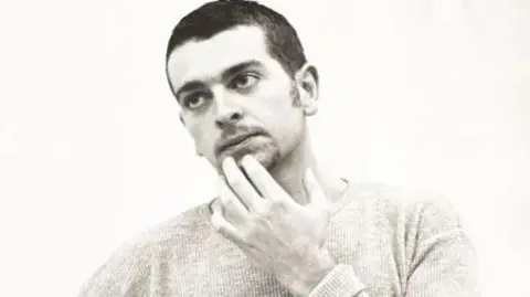 Geoff Hopson A black and white photo of a younger Geoff Hopson who has dark short hair and has his fingers on his chin looking thoughtful.