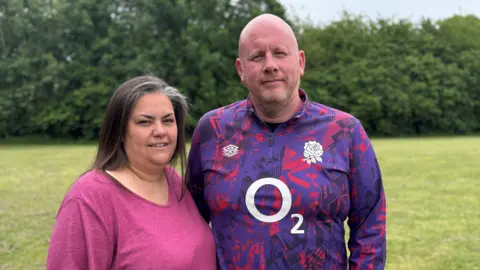 A woman and a man are standing side by side in a field with trees behind them. The woman is wearing a pink top and has brown hair with some grey strands at the front. The man is wearing a pink and purple patterned rugby top. He is bald with a short beard.