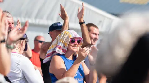 Kirstin McEwan A woman with pink sunglasses and a towel on her head to help cope with the heat claps in the middle of a crowd