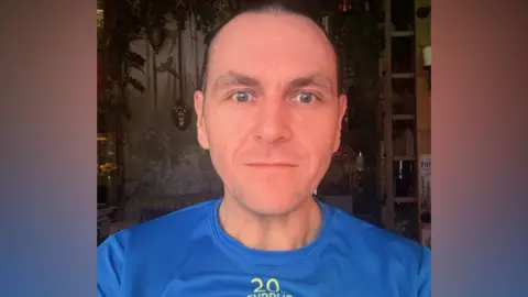 David Stone A man wearing a blue T-shirt with a yellow logo on it looks at the camera.