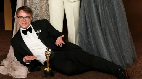 A man in a tuxedo lies on the floor in front of a golden Oscar statuette 