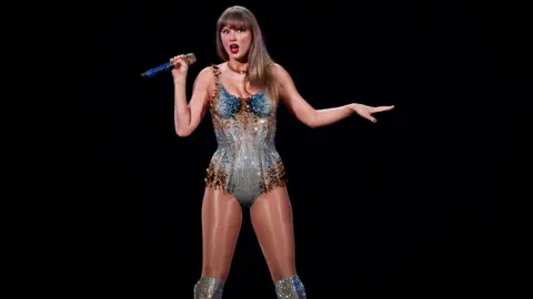 REUTERS/Jennifer Gauthier Taylor Swift stands against a black background in a sparkling blue and borwn corseted bodysuit. She is holding a mic and has her arms pushed out from her sides. 