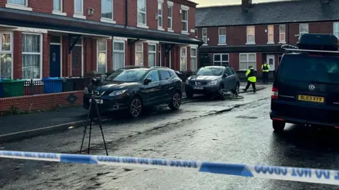 BBC News/Molly Brewer Police cordon strapped across street with two police officers and a tripod seen near parked cars outside terraced houses
