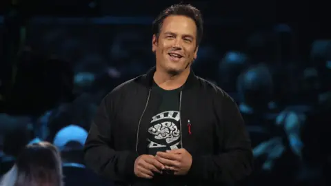 Getty Images A man wearing a bomber jacket over a black t-shirt with white print stands on stage addressing a crowd