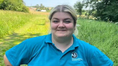 Megan is smiling at the camera, she is wearing a blue polo shirt which has a badger logo and reads Gloucestershire Wildlife Trust in white writing.