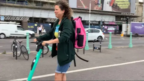 A woman, Shona Mackintosh, riding a turquoise e-scooter on a seafront cycle lane