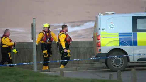 Darrin Stevens Three RNLI volunteers dressed in yellow and black and wearing life jackets walking past a parked police van. 