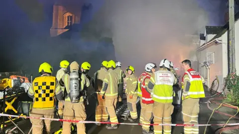 Stuart Howells/BBC Twelve firefighters standing on the other side of red and white tape with smoke billowing in front of them. They all have light brown uniform on with helmets and hi-vis. One is wearing breathing apparatus on their back.