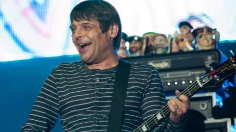 PA Media Mani on stage with short brown slightly greying hair playing his base guitar wearing a horizontal-striped sweatshirt at the V Festival in Stafford in 2012.