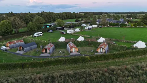 Katryna Shell An aerial view of a campsite, with yurts and cabins with campervans in the distance. 