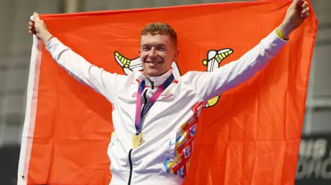 Reuters. Matthew lleva una chaqueta blanca de los Juegos de la Commonwealth con la medalla alrededor del cuello. Sostiene una gran bandera roja de la Isla de Man detrás de él y sonríe.