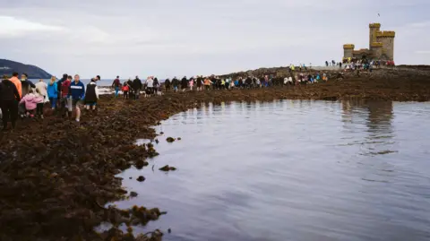 Douglas City Centre Management Large groups of people walking over seaweed to a small tower on an island