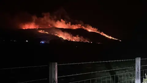 Claire Paine The image shows a large fire burning in the dark at the foot hill of the Mourne Mountains in Castlewellan. A small, barbed wire fence is illuminated in the foreground.