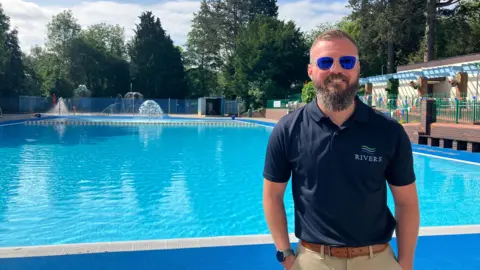 BBC Droitwich Spa Lido manager Seb Parker stands by the pool on a sunny day with his sunglasses on. The lido is bright blue in the sunlight and there are water features in the background.
