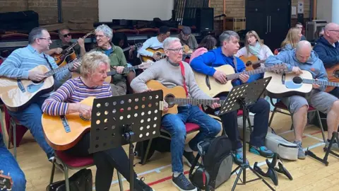 An ensemble of guitarists of all ages sit in two lines at a club with music stands in front of some of them.