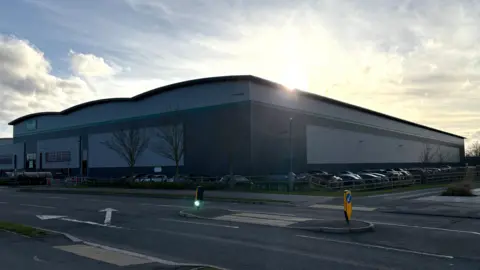 Ben Schofield/BBC A large warehouse, almost in silhouette, with the sun about to dip below its roofline. The warehouse is on the other side of a well-kept road and is surrounded by a car park, which is full of cars. There are wispy clouds in the sky. 