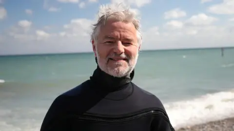 Shaun Whitmore/BBC Diver Rob Spray is standing on a pebble beach in a black wet suit. Behind him is the blue water of the sea crashing against the shoreline. He is looking directly at the camera and smiling.