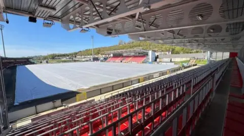 A number of seats are visible at the Ryan McBride Brandywell Stadium.
A large white tarpaulin sheet is covering the football pitch. 