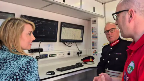 Northamptonshire Search and Rescue Interior of a control room inside a mobile or modular unit, with two wall-mounted computer screens displaying search-and-rescue software, including a digital map with marked locations and a status dashboard. Two men and a woman are in vision.
