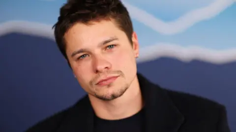 Toby is looking into the camera, he has a black t-shirt on with a black jacket over the top. He has short brown hair and blue eyes.
