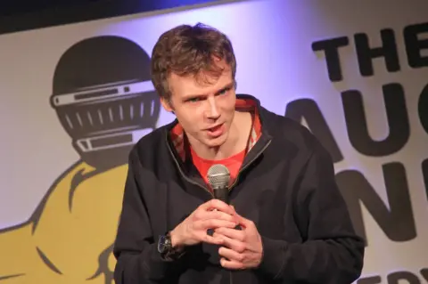 Jack Kelly Tom Little performing stand-up comedy. He is holding a microphone onstage anis talking to the audience. He has short brown hair and blue eyes. He wears a blue zip-up sweater over a red t-shirt.