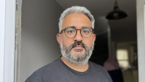 Aman is stood in the centre of the frame, he is wearing round glasses and has grey hair with a grey beard and a grey t-shirt