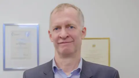 Imperial College London Professor Eric Yeatman, a man wearing a blue shirt and dark suit jacket is standing in a room with a plain white wall which has framed certificates hanging on it.