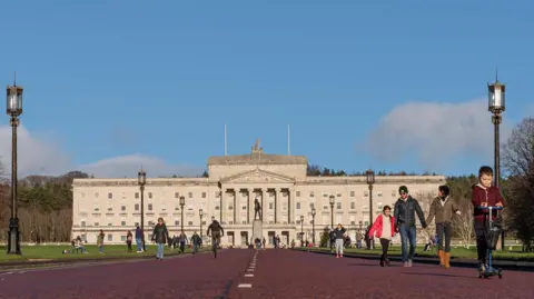 Getty Images Numerous people walking, scooting, cycling and playing in front of Stormont. It is a large stone building with stairs leading up to it. It is rectangular shape and has four rows of windows along the front, and a stone turrets at the front. On top of the building is a metal statue. Dotted around the grounds are black lampposts and benches for people to sit on. There is a statue feature in front of the steps to the front door.