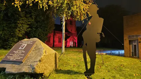 A thin, black metal silhouette statue of a soldier leaning on a rifle is held up on grass by four strings. It stands next to a large granite stone with a memorial plaque on it. There is a red poppy on the statue. In the background, the church is illuminated by red floodlights.