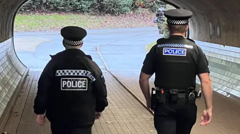 Two police officers walking through an underpass and away from the camera; they have their backs to us. Both of them are in uniform. The officer on the left is slightly shorter and is wearing a dark police officer's uniform, with a long-sleeved jacket and a peaked cap. The officer on the right is taller, is also wearing a uniform, though he is wearing a t-shirt and his bare lower arms can be seen. The pair are walking on paving stones towards the bright tunnel exit. The walls are curved, with the lower section covered with shiny tiles.