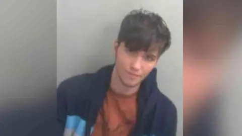 Essex Police Chase Wiseman has short brown hair with a fringe in his police custody photo. He has a blue jacket and a camel brown T-shirt on.